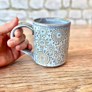 May include: A handmade ceramic mug with a light blue glaze and a textured pattern of circles and dots. The mug has a curved handle and is held by a hand. The mug is on a wooden surface.