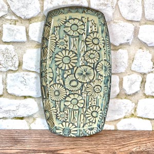 May include: A rectangular ceramic tray featuring a light green glaze with a dark, recessed floral pattern. The design consists of multiple stylized daisy-like flowers and vertical stems covering the entire surface, finished with a dark rim.