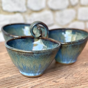 May include: A handmade ceramic serving dish with three connected bowls. The bowls have a blue and green glaze with a glossy finish. The handle is a twisted loop. The dish is on a wooden surface.