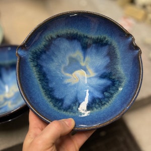 Puede incluir: Un cuenco de cer&aacute;mica hecho a mano con un esmalte azul oscuro y un centro azul claro y amarillo. El cuenco tiene una forma &uacute;nica con dos peque&ntilde;as asas a los lados y un borde marr&oacute;n. El cuenco se utiliza probablemente para servir alimentos.
