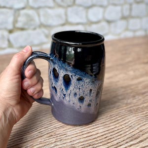 May include: A handcrafted ceramic mug with a large handle. The mug features a glossy black rim and a gradient design, transitioning from dark blue to a lighter, speckled blue and purple. The mug is held by a hand.