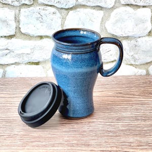 May include: A tall, tapered ceramic travel mug with a speckled blue glaze and a dark brown rim. The mug features a sturdy side handle and comes with a removable black silicone lid that has the text CAUTION VERY HOT embossed on the top surface.