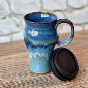 May include: A handmade ceramic mug with a blue and turquoise glaze, featuring a unique handle and a matching black lid. The mug has a capacity of approximately 12 ounces and is suitable for hot or cold beverages.