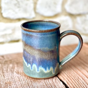 May include: A handmade ceramic mug with a blue and brown glaze. The mug has a rounded handle and a brown rim. The colors blend together, creating a unique, artistic look. The mug is perfect for coffee, tea, or any other beverage.