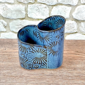 May include: A dual-compartment ceramic utensil holder featuring a vibrant blue glaze with a dark, recessed floral or sunburst pattern embossed across the exterior surface. The vessel has two distinct cylindrical openings of varying heights.