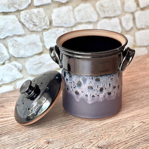 May include: A cylindrical ceramic crock with two side handles and a matching lid. The vessel features a glossy black upper section that transitions into a textured, dripped white and lavender glaze over a matte purple base, with lid vents.
