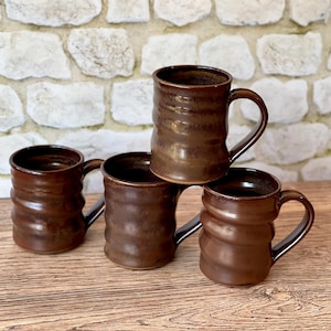 May include: Four brown ceramic mugs with a textured, ridged design. Each mug has a curved handle and a glossy finish. The mugs are stacked on a wooden surface, with a stone wall in the background. The mugs are a warm, earthy tone.