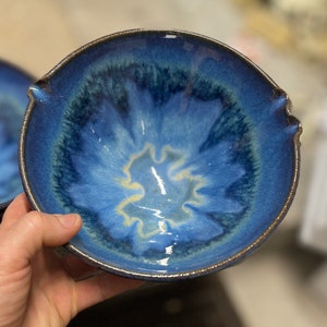 Puede incluir: Un cuenco de cer&aacute;mica hecho a mano con un vibrante esmalte azul. El cuenco presenta un dise&ntilde;o &uacute;nico con un centro m&aacute;s claro, casi amarillo, y bordes azul oscuro. El borde tiene un sutil ribete marr&oacute;n dorado, a&ntilde;adiendo un toque de elegancia.