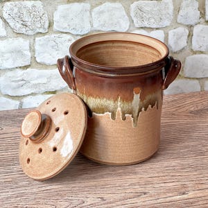 May include: A cylindrical ceramic crock with two side handles and a matching lid featuring multiple ventilation holes. The vessel displays a two-tone glaze with a dark brown upper section transitioning into a lighter tan base with drip patterns.