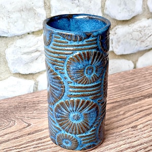 May include: A cylindrical ceramic tumbler featuring a deep blue glaze with a textured, carved pattern of dark brown circular floral motifs and horizontal lines. The vessel has a smooth, open top and a straight-sided body with a visible clay base.