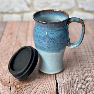 May include: A ceramic travel mug featuring a two-tone blue glaze with a darker blue upper section and a lighter blue base. The mug has a curved handle and a dark brown rim. A separate, circular black silicone lid sits beside the mug on the surface.