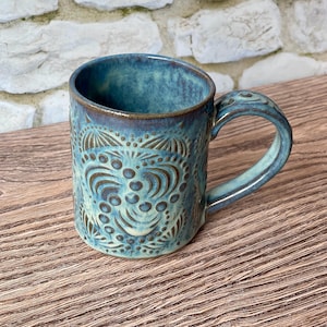 May include: A ceramic mug featuring a variegated blue and teal glaze with a dark brown rim. The exterior surface is decorated with an embossed pattern of repeating circular dots and curved lines that create a textured, symmetrical design.