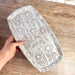 May include: A rectangular ceramic tray with a white glaze and a debossed floral pattern featuring multiple daisy-like flowers. The recessed areas of the floral design are highlighted in a dark brown color, creating a high-contrast texture.