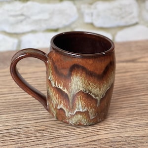 May include: A handmade ceramic mug with a brown interior and a textured exterior. The mug features a unique glaze with shades of brown, tan, and cream, creating a layered, organic pattern. The mug has a rounded body and a sturdy handle.