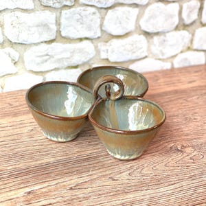 May include: A three-section ceramic serving dish with a handle. The dish has a mottled glaze in shades of green and brown, with a darker brown rim. The handle is a loop shape. The dish is on a wooden surface.