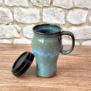 May include: A handmade ceramic travel mug with a teal and blue glaze. The mug has a handle and a black lid. The mug is on a wooden surface.