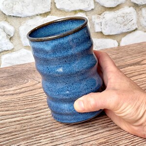 May include: A cylindrical ceramic tumbler with a wavy, rippled exterior surface. The vessel features a deep blue speckled glaze with a dark brown rim. The texture of the cup is uneven, creating a series of horizontal ridges along the body.