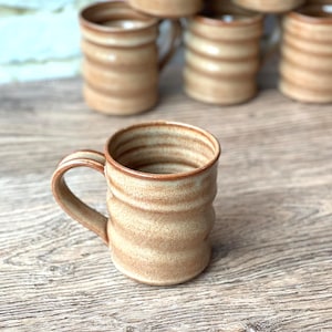 May include: A handmade ceramic mug with a tan and brown color scheme. The mug has a ribbed design and a curved handle. Several similar mugs are stacked in the background. The mugs are approximately 4 inches tall.