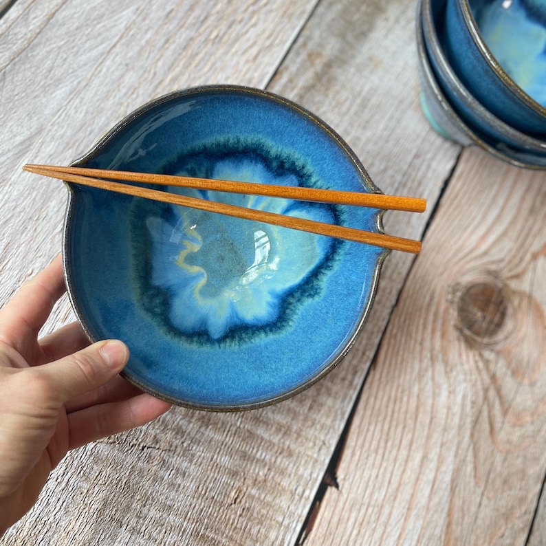 Puede incluir: Un cuenco de cer&aacute;mica azul hecho a mano con un dise&ntilde;o &uacute;nico, con un centro azul claro y bordes azul oscuro. Dos palillos de madera descansan sobre el cuenco. El cuenco forma parte de un juego, con otros visibles en el fondo.