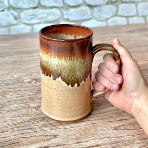 May include: A tall ceramic mug featuring a reactive glaze with a deep brown rim that transitions into a lighter tan and beige speckled base. The mug has a cylindrical body and a curved handle, showcasing a distinct drip pattern where the dark glaze meets the lighter clay body.