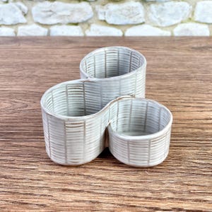 May include: A ceramic organizer featuring three interconnected cylindrical compartments of varying heights. The exterior and interior surfaces are finished in a light off-white glaze with a repeating pattern of thin, dark brown vertical and horizontal lines.