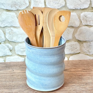 May include: A light blue ceramic utensil holder with a wavy, cylindrical body containing a collection of various wooden kitchen utensils, including a slotted spoon, a solid spoon, and a spatula with a circular cutout in the handle.