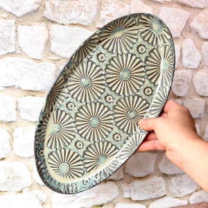 May include: An oval ceramic serving platter featuring a repeating embossed floral pattern of stylized daisies in shades of sage green and cream. The platter has a dark, glazed rim and a glossy finish, showcasing a detailed, textured surface design.