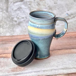 May include: A ceramic travel mug with a glossy glaze featuring blended shades of blue, teal, and a horizontal band of yellow. The mug has a sturdy handle and comes with a removable black plastic lid that has the text CAUTION: VERY HOT.