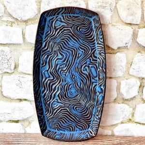 May include: A rectangular ceramic tray featuring a deep blue glaze with a raised, dark brown topographic line pattern. The surface has a textured, wavy design that mimics contour lines, with a smooth, dark brown rim framing the entire piece.
