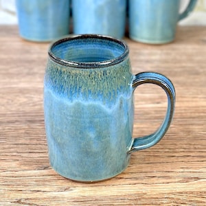 May include: A ceramic mug with a blue and green glaze. The mug has a large handle and a dark brown rim. Several other mugs are visible in the background, all with a similar design. The mug is on a wooden surface.