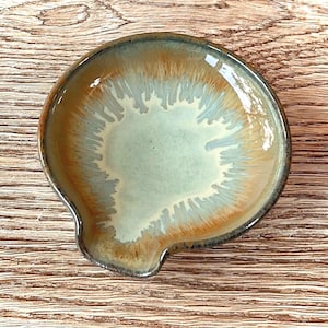 May include: A ceramic spoon rest with a unique glaze. The center is a pale green, surrounded by a blend of brown and tan hues. The rim is a darker brown. The rest has a small, curved handle.
