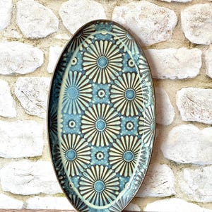 May include: An oval ceramic platter featuring a repeating pattern of dark brown and light blue stylized flower motifs. The dish has a dark brown rim and a glazed finish, with the floral design arranged in a grid across the entire surface.