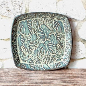 May include: A square ceramic plate featuring a relief pattern of overlapping monstera leaves. The surface displays a dual-tone finish with dark blue-green recessed areas and a lighter, creamy off-white glaze on the raised leaf textures.