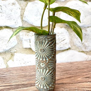 May include: A cylindrical ceramic vase featuring a relief pattern of stylized sunbursts or flowers in shades of sage green and dark brown, containing a green artificial plant with variegated leaves extending from the top opening.