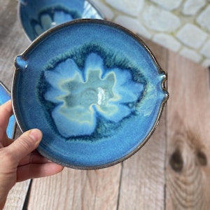 Puede incluir: Un juego de tres cuencos de cer&aacute;mica hechos a mano con un esmalte azul brillante. Cada cuenco presenta un dise&ntilde;o floral &uacute;nico en el centro, con una paleta de colores azul claro y crema. Los cuencos tienen peque&ntilde;as asas a cada lado.