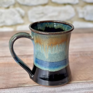 May include: A handmade ceramic mug with a curved handle. The mug features a gradient of colors, including black at the base, transitioning to blue, turquoise, and a tan-orange near the rim. The interior is a deep blue.