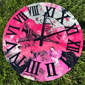 May include: A round wall clock with a pink, white, and black abstract design. The clock face features black Roman numerals, the Eiffel Tower, and the word "Paris". Butterflies and the phrase "Ooh la la" are also included.