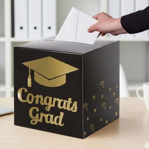 May include: A black square card box featuring a gold graduation cap icon and the text Congrats Grad in gold script lettering. The side of the box is decorated with small gold stars and swirl patterns, with a slot on top for inserting white envelopes.