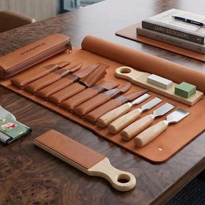 May include: A brown leather tool roll containing several wood carving chisels with wooden handles and protective leather covers. The set includes a matching pencil case labeled SONGMICS, a sharpening stone block, a leather strop, and a gray glove.