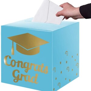 May include: A light blue square card box featuring a gold foil graduation cap icon and the text "Congrats Grad" in gold script. The side of the box is decorated with small gold stars and swirls. A white envelope is being inserted into the top slot.