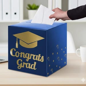 May include: A square blue card box featuring a gold graduation cap icon and the text "Congrats Grad" in gold lettering. The box has a slot on the top for inserting envelopes and is decorated with small gold star and swirl patterns on the side.