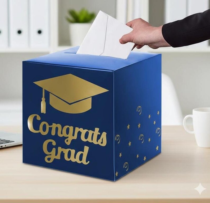 May include: A square blue card box featuring a gold graduation cap icon and the text "Congrats Grad" in gold lettering. The box has a slot on the top for inserting envelopes and is decorated with small gold star and swirl patterns on the side.