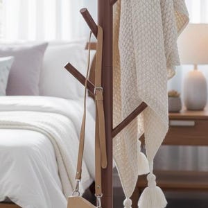 May include: A brown wooden coat rack with a central pole and multiple angled hooks. A straw hat with a black band hangs from one hook, a beige crossbody bag hangs from another, and a cream-colored textured throw blanket with tassels is draped over it.