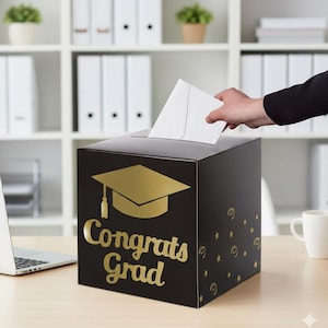 May include: A black graduation card box with gold lettering that says "Congrats Grad" and a gold graduation cap design. A hand is placing a white envelope into the slot on top of the box. The box is on a wooden desk.