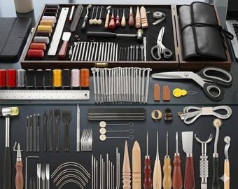 May include: A comprehensive leather crafting tool kit featuring a wooden storage box, metal rulers, various needles, thread spools in multiple colours, a hammer, scissors, awls, punches, burnishers, and specialised cutting blades arranged on a flat surface.