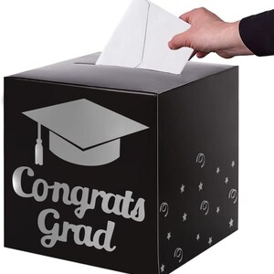 May include: A black square card box featuring a silver graduation cap icon and the text "Congrats Grad" in a script font. The side of the box is decorated with silver stars and swirls. A white envelope is being inserted into the top slot.
