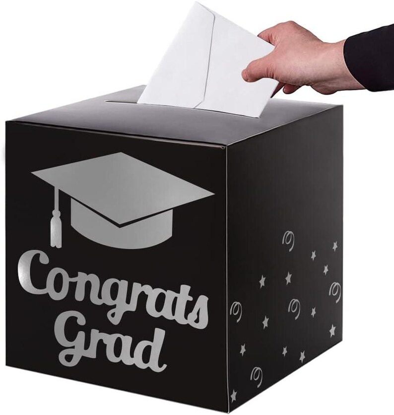 May include: A black square card box featuring a silver graduation cap icon and the text "Congrats Grad" in a script font. The side of the box is decorated with silver stars and swirls. A white envelope is being inserted into the top slot.