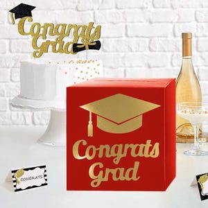 May include: A red square card box featuring a gold foil graduation cap icon and the text Congrats Grad. The set includes a white cake with a gold glitter Congrats Grad topper and two small white place cards with black borders and gold accents.