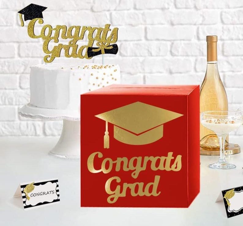 May include: A red square card box featuring a gold foil graduation cap icon and the text Congrats Grad. The set includes a white cake with a gold glitter Congrats Grad topper and two small white place cards with black borders and gold accents.