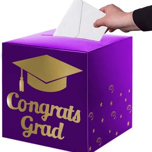 May include: A square purple card box featuring a gold graduation cap icon and the text "Congrats Grad" in gold lettering. The box has a slot on the top for inserting cards, and the sides are decorated with small gold stars and swirl patterns.